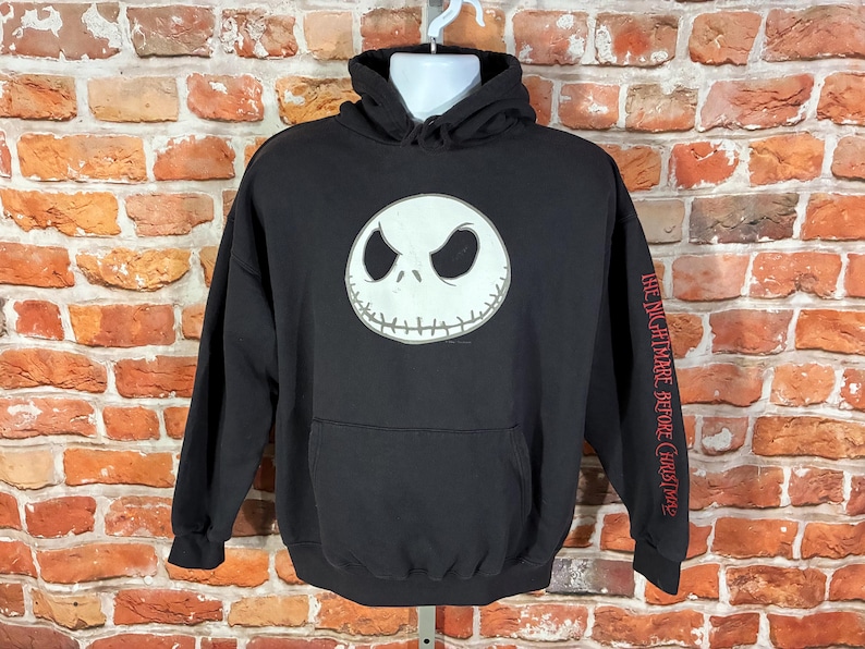 May include: Black hooded sweatshirt featuring a white Jack Skellington face on the front. The sleeve has the text "THE NIGHTMARE BEFORE CHRISTMAS" in red. The sweatshirt has a front pocket and a drawstring hood.