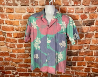 May include: A short-sleeved button-down shirt with a floral pattern. The shirt features a pink, teal, and lavender color scheme with white flower accents. The shirt has a pocket on the left side and is displayed on a mannequin.