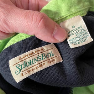 May include: A black and green polo shirt with a green collar and a black label that says "AUTHENTIC STJOHNSBAY ESTABLISHED 1984". A white label with the text "M MEDIANO 100% COTTON/ ALGODON MADE IN HONG KONG HECHO EN HONG KONG RN # 93677" is visible.