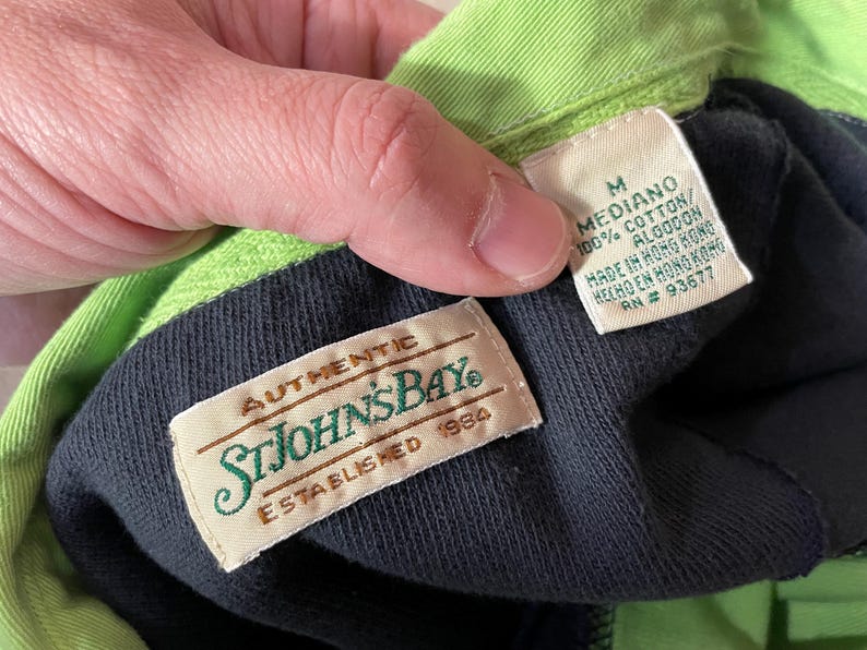 May include: A black and green polo shirt with a green collar and a black label that says "AUTHENTIC STJOHNSBAY ESTABLISHED 1984". A white label with the text "M MEDIANO 100% COTTON/ ALGODON MADE IN HONG KONG HECHO EN HONG KONG RN # 93677" is visible.