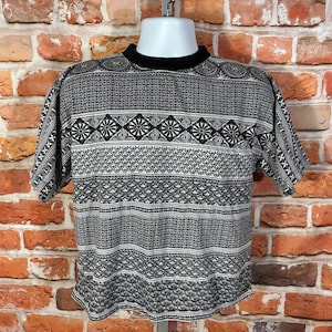 May include: A short-sleeve t-shirt featuring a black and white geometric pattern with horizontal bands of varying designs, including diamonds and floral motifs. The garment has a solid black crew neckline and a straight hemline throughout.