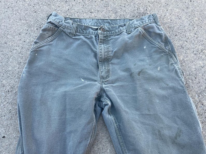 Puede incluir: Un par de pantalones de trabajo verde claro con salpicaduras de pintura blanca. Los pantalones tienen un dise&ntilde;o tradicional de cinco bolsillos y cierre de bot&oacute;n.