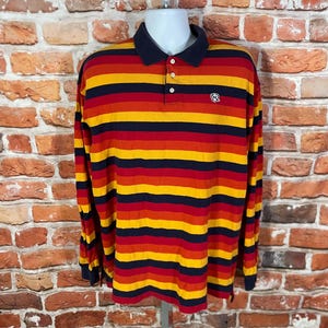 May include: A long-sleeved polo shirt with horizontal stripes in red, yellow, and navy blue. The shirt has a navy blue collar and cuffs, with a small logo on the chest. The shirt is displayed on a mannequin.