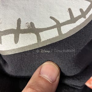 May include: Close-up of a black garment featuring a white and gray graphic of a character's face. The graphic has a stitched appearance. The text "&copy; Disney / Touchstone" is visible.
