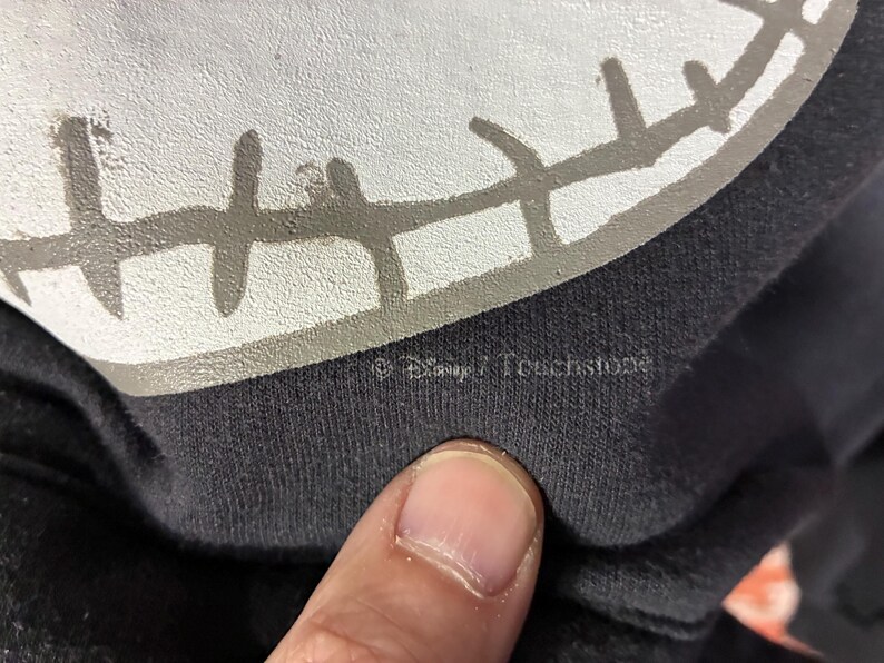 May include: Close-up of a black garment featuring a white and gray graphic of a character's face. The graphic has a stitched appearance. The text "&copy; Disney / Touchstone" is visible.