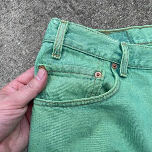 May include: Close-up of a pair of green denim jeans. The jeans have a classic five-pocket design with copper rivets and a button closure. The fabric has a slightly faded appearance.