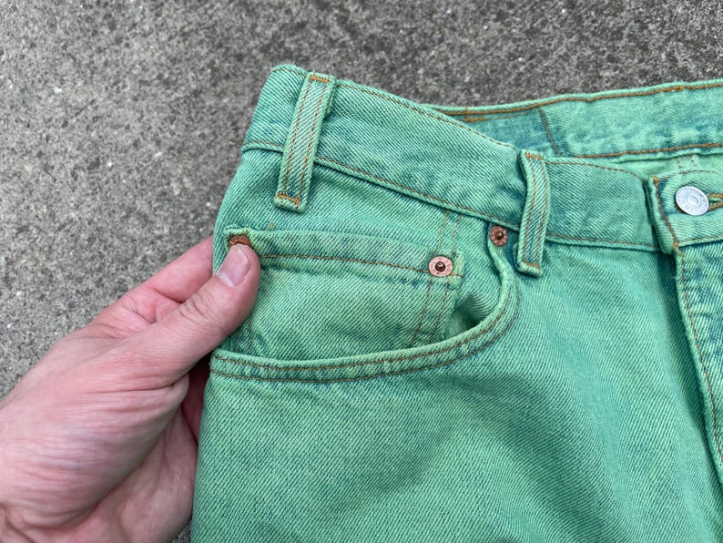 May include: Close-up of a pair of green denim jeans. The jeans have a classic five-pocket design with copper rivets and a button closure. The fabric has a slightly faded appearance.