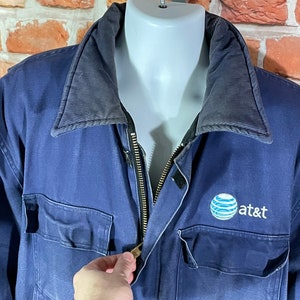 May include: A blue work jacket with a black zipper and a navy blue collar. The jacket has a large AT&T logo on the right chest.