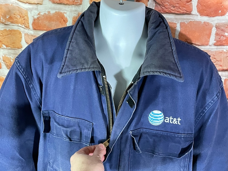 May include: A blue work jacket with a black zipper and a navy blue collar. The jacket has a large AT&T logo on the right chest.