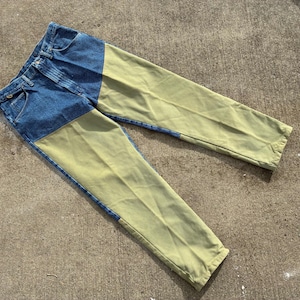May include: Blue denim jeans with olive green panels on the front of the legs. The jeans are laid flat on a concrete surface, showcasing the contrasting colors and design. The jeans have a classic five-pocket design.