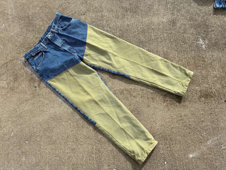 May include: Blue denim jeans with olive green panels on the front of the legs. The jeans are laid flat on a concrete surface, showcasing the contrasting colors and design. The jeans have a classic five-pocket design.