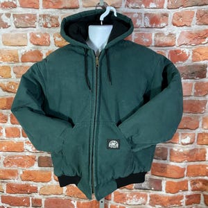 May include: A dark green hooded jacket with a black interior lining and a full-length zipper. The jacket has two front pockets and a black ribbed hem and cuffs. A small patch is visible on the front pocket.