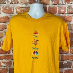 May include: A yellow t-shirt with embroidered graphics of a rainbow, sun, and the NBC peacock logo. The text "The Today Show" is embroidered below the graphics.