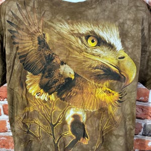 Puede incluir: Camiseta marr&oacute;n con un gr&aacute;fico detallado de un &aacute;guila. El dise&ntilde;o muestra un &aacute;guila con las alas extendidas, un primer plano de su cabeza y un &aacute;guila m&aacute;s peque&ntilde;a en una rama. La camiseta tiene un efecto tie-dye.