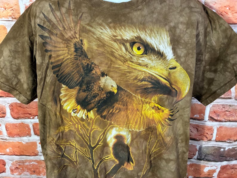Puede incluir: Camiseta marr&oacute;n con un gr&aacute;fico detallado de un &aacute;guila. El dise&ntilde;o muestra un &aacute;guila con las alas extendidas, un primer plano de su cabeza y un &aacute;guila m&aacute;s peque&ntilde;a en una rama. La camiseta tiene un efecto tie-dye.