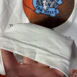 May include: White t-shirt featuring a basketball design with the words "Tar Heels" in blue and a basketball graphic. The shirt is made of a soft, light-colored fabric.