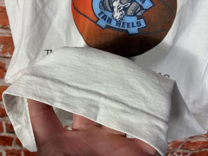May include: White t-shirt featuring a basketball design with the words "Tar Heels" in blue and a basketball graphic. The shirt is made of a soft, light-colored fabric.