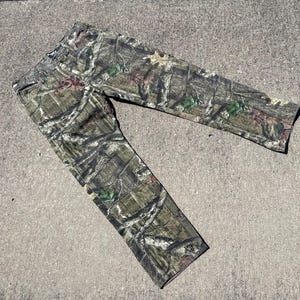 May include: Camouflage pants in a woodland pattern, featuring shades of green, brown, and tan. The pants are made of a durable fabric and are designed for outdoor activities. The pants are laid flat on a concrete surface.