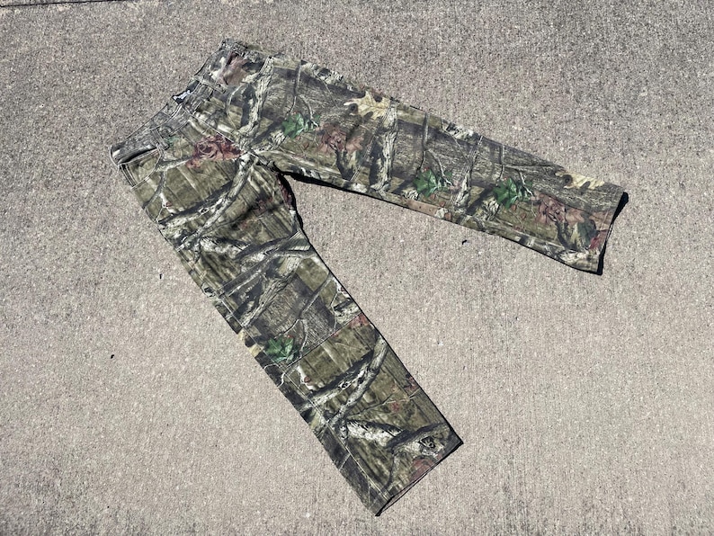 May include: Camouflage pants in a woodland pattern, featuring shades of green, brown, and tan. The pants are made of a durable fabric and are designed for outdoor activities. The pants are laid flat on a concrete surface.