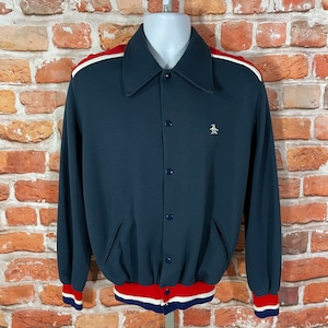 Puede incluir: Una chaqueta azul marino con botones, con rayas rojas y blancas en los hombros y los puños. La chaqueta tiene un pequeño logotipo de pingüino bordado en el pecho.