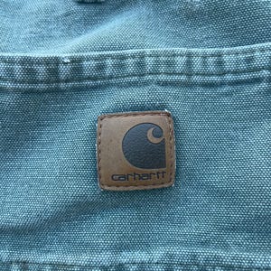 Puede incluir: Primer plano de un bolsillo de lona azul con un parche de cuero marr&oacute;n Carhartt. El parche tiene un logotipo de luna creciente blanco y la palabra "Carhartt" en letras blancas.