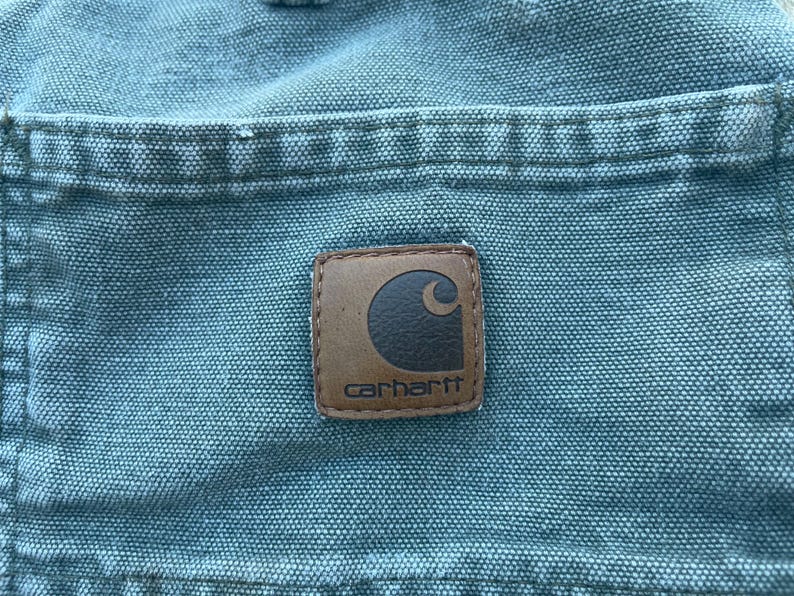 Puede incluir: Primer plano de un bolsillo de lona azul con un parche de cuero marr&oacute;n Carhartt. El parche tiene un logotipo de luna creciente blanco y la palabra "Carhartt" en letras blancas.