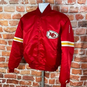 May include: A red satin bomber jacket with white and yellow stripes on the sleeves. The jacket features a team logo patch on the chest. The jacket is displayed on a mannequin against a brick wall.