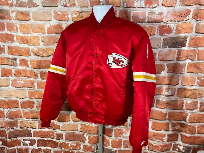 May include: A red satin bomber jacket with white and yellow stripes on the sleeves. The jacket features a team logo patch on the chest. The jacket is displayed on a mannequin against a brick wall.