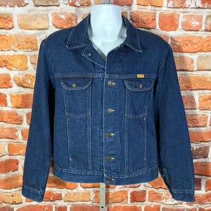 May include: A dark blue denim jacket with a button-up front, two chest pockets, and two side pockets. The jacket has a collar and long sleeves. A small yellow tag is visible on the chest pocket.