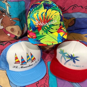 May include: Three vintage trucker hats with different designs. The first hat is green, yellow, and blue with the text "Bahamas" embroidered on the front. The second hat is blue and white with the text "St. Maarten, N.I." and a sailboat graphic. The third hat is red and white with the text "St. Thomas, V.I." and a palm tree graphic.