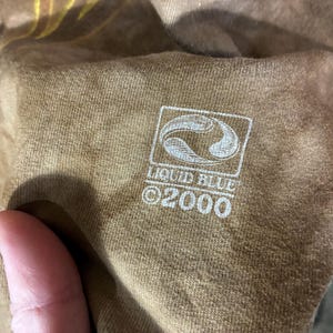 Puede incluir: Primer plano de una camiseta marr&oacute;n con el logotipo blanco de Liquid Blue y el texto "&copy;2000". El logotipo es un dise&ntilde;o estilizado dentro de un marco cuadrado.