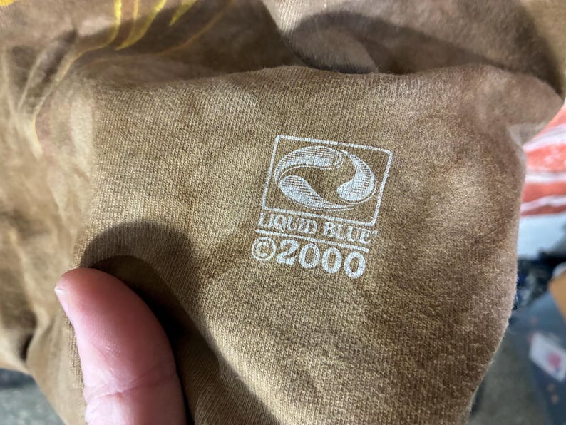 Puede incluir: Primer plano de una camiseta marr&oacute;n con el logotipo blanco de Liquid Blue y el texto "&copy;2000". El logotipo es un dise&ntilde;o estilizado dentro de un marco cuadrado.