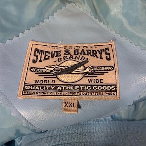 May include: Close-up of a light blue jacket interior with a stitched label. The label reads "STEVE & BARRY'S BRAND" with an airplane graphic and the text "WORLD WIDE QUALITY ATHLETIC GOODS". The size tag reads "XXL".