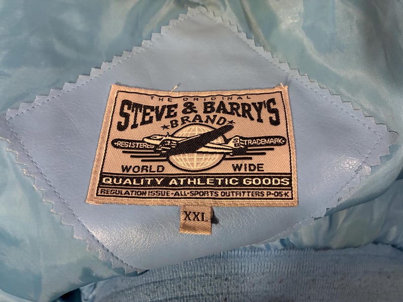 May include: Close-up of a light blue jacket interior with a stitched label. The label reads "STEVE & BARRY'S BRAND" with an airplane graphic and the text "WORLD WIDE QUALITY ATHLETIC GOODS". The size tag reads "XXL".
