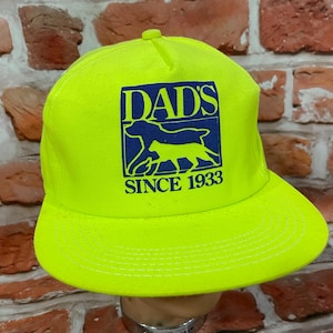 May include: Bright yellow baseball cap with a flat brim. The cap features a blue and yellow graphic with the text "DAD'S" above a dog silhouette and the text "SINCE 1933".