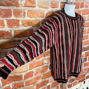 Peut inclure: Un pull &agrave; manches longues avec un motif ray&eacute; noir, rouge, blanc et gris. Le pull a un col rond et est fait d'un tissu doux et tricot&eacute;.