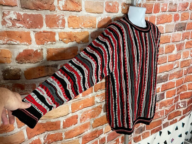 Peut inclure: Un pull &agrave; manches longues avec un motif ray&eacute; noir, rouge, blanc et gris. Le pull a un col rond et est fait d'un tissu doux et tricot&eacute;.