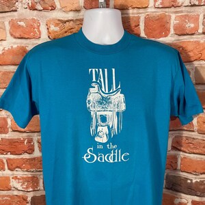 May include: A turquoise blue t-shirt with a white graphic of a saddle and the text "Tall in the Saddle".
