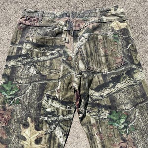 May include: Camouflage-patterned pants in shades of green, brown, and tan. The pants feature two back pockets and a classic five-pocket design. The fabric has a detailed print resembling a forest environment.