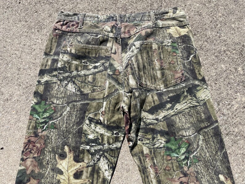 May include: Camouflage-patterned pants in shades of green, brown, and tan. The pants feature two back pockets and a classic five-pocket design. The fabric has a detailed print resembling a forest environment.