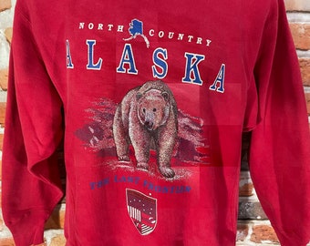 May include: Red sweatshirt with a graphic of a brown bear and the text "NORTH COUNTRY ALASKA THE LAST FRONTIER" in blue. The graphic also includes a small outline of Alaska.