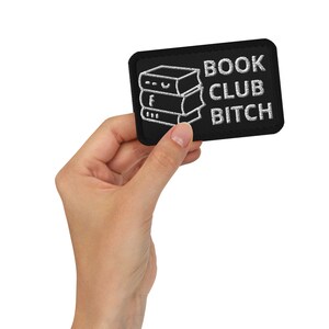 Puede incluir: Parche bordado negro con las palabras "BOOK CLUB BITCH" en blanco, junto con una pila de tres libros. El parche es rectangular con esquinas redondeadas.