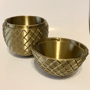 May include: A gold-colored, 3D printed, egg-shaped container with a textured, diamond-shaped pattern. The container is open, revealing the inside of the egg.