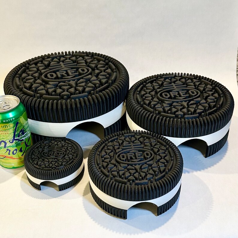May include: Four black Oreo cookie-shaped cake stands of varying sizes. The largest stand is in the back, and the smallest stand is in the front. The stands are all made of black cake with a white rim.
