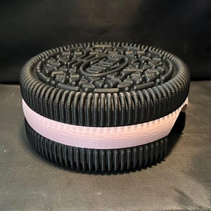 May include: A black and white cookie-shaped container with a pink stripe around the middle. The cookie has a raised design on the top.