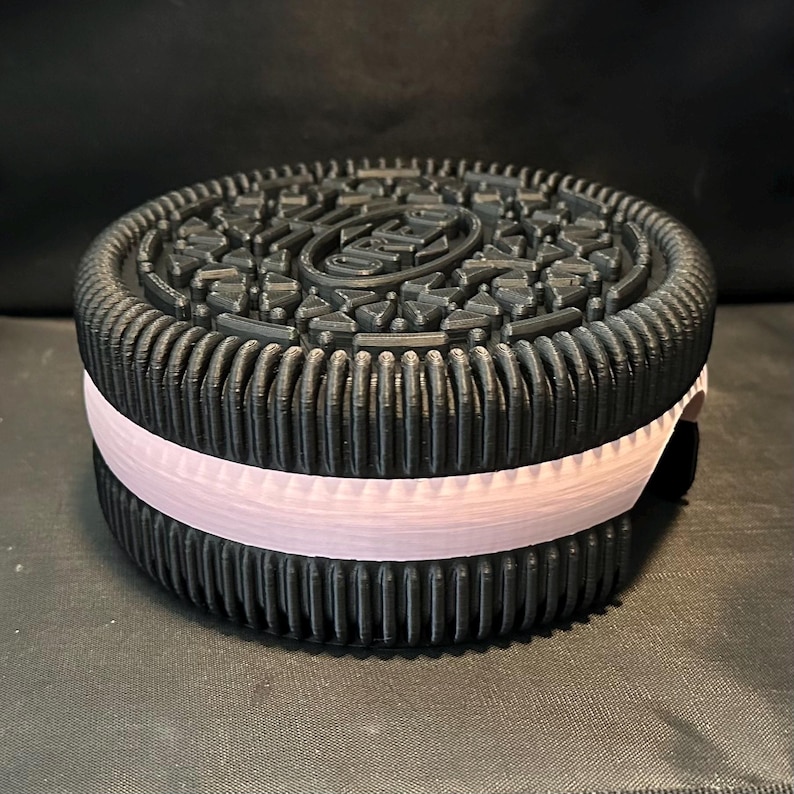 May include: A black and white cookie-shaped container with a pink stripe around the middle. The cookie has a raised design on the top.