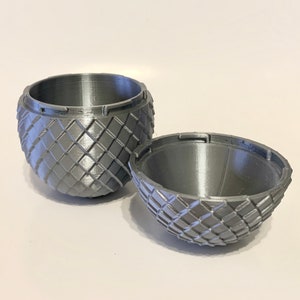 May include: A silver, 3D printed, egg-shaped container with a textured, diamond-shaped pattern. The container is open, revealing the inside of the egg.