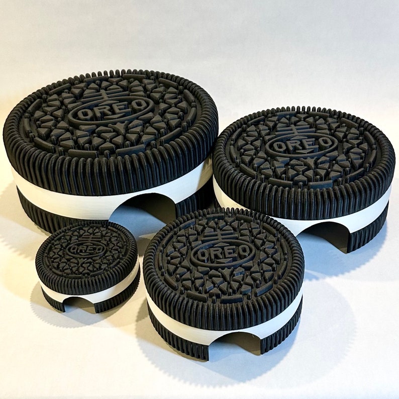 May include: Four black and white Oreo cookie-shaped cake stands, the largest is at the top and the smallest is at the bottom. The stands are all the same design, with a white band around the middle and the Oreo logo on the top.