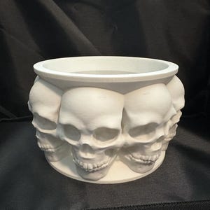 May include: White ceramic planter shaped like a circle of skulls. The planter is made of smooth, white ceramic and features a ring of skulls around the top. The skulls are all facing forward and have a detailed design.