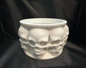 May include: White ceramic planter shaped like a circle of skulls. The planter is made of smooth, white ceramic and features a ring of skulls around the top. The skulls are all facing forward and have a detailed design.